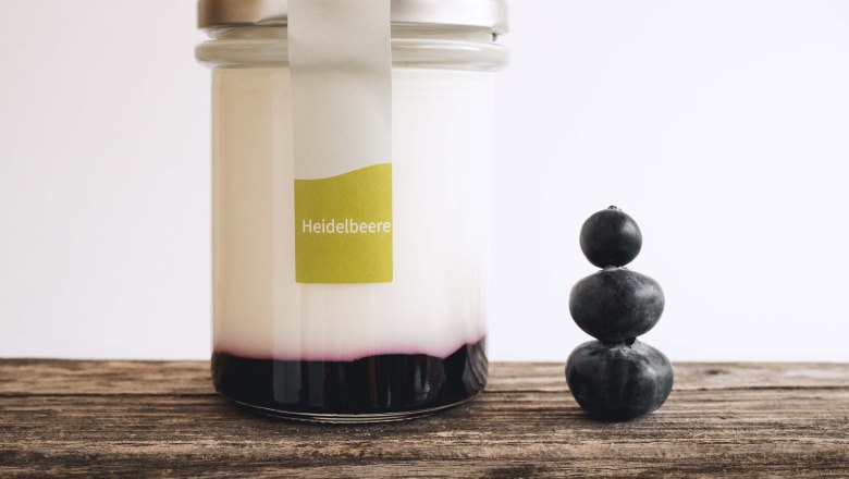 Yogurt organic farm at Stickelberg, © Anni Lechner Fotografie A glass filled with yogurt and fruit on a wooden table, next to it 3 stacked blueberries.
