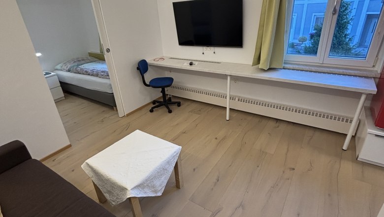TDR House Morning Sun Living Room, © Fam Pauker Modern living room with brown sofa, small table, TV on the wall, desk and chair. Window with a view of a blue building.