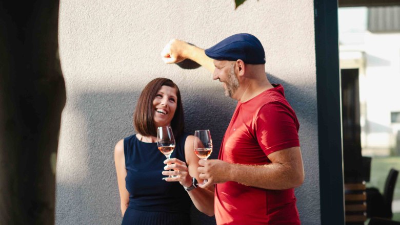 Sigrid and Christoph Schleinzer, © Astrid Bartl A man and a woman stand smiling against a wall, holding glasses of wine and shaded by leaves.