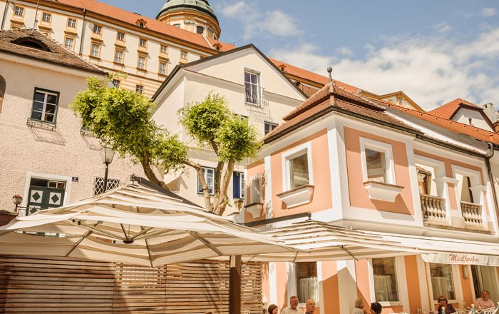 Café-Confectionery Mistlbacher, © Best of Wachau/Rita Newman Outdoor area of a café with parasols and guests, in the background a historic building with a dome.