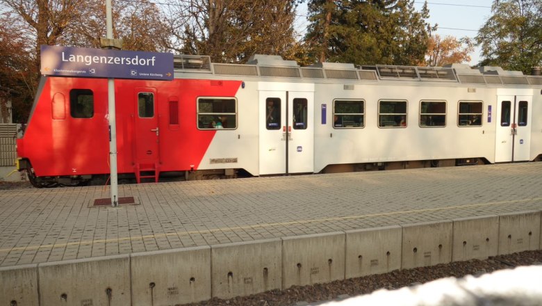 Langenzersdorf train station, © Leader-Region Weinviertel / Lahofer A train stands at Langenzersdorf station, with a sign in the foreground. The train is red and white and the surrounding area is surrounded by trees.