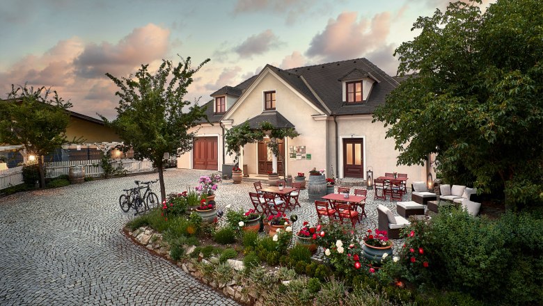 Cozy winegrower's terrace, © Martin Sommer A cozy courtyard with tables, chairs and flower beds in front of a large house at sunset.