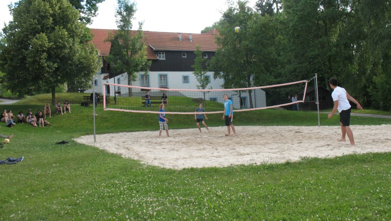 Theater and vacation village Königsleitn, © Theater- und Feriendorf Königsleitn People playing volleyball on an outdoor sand court, surrounded by trees and a building in the background.