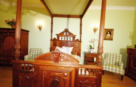 Renaissancehof Kerschner, © Renaissancehof Kerschner An elegant bedroom with an ornately carved wooden four-poster bed, green checkered armchairs and antique furniture.