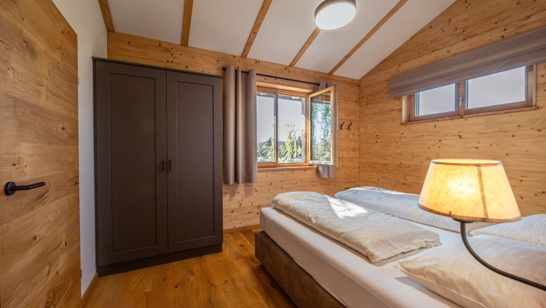 Kittenberger Chalets at the Gartensee, © Niederösterreich Werbung / Maximilian Pawlikowsky Interior view of a bedroom in a chalet with wooden walls, a double bed, a closet and a window with a view outside.