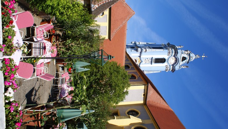 Flower garden, © Schendl A flowering garden with pink chairs and tables in front of a historic building with a blue tower.