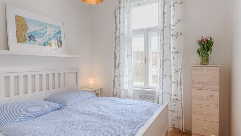 Top 3 - Bedroom, © VP FeWo OG Bright bedroom with white bed, blue bed linen, wooden chest of drawers and window with curtains.