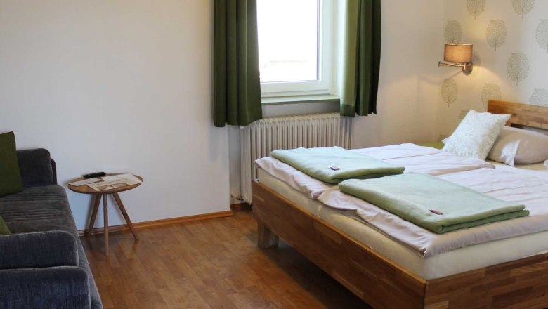 Double room, © Magdalena Donhauser Bedroom with double bed, window with green curtains, sofa, table, wallpaper and a wall lamp.