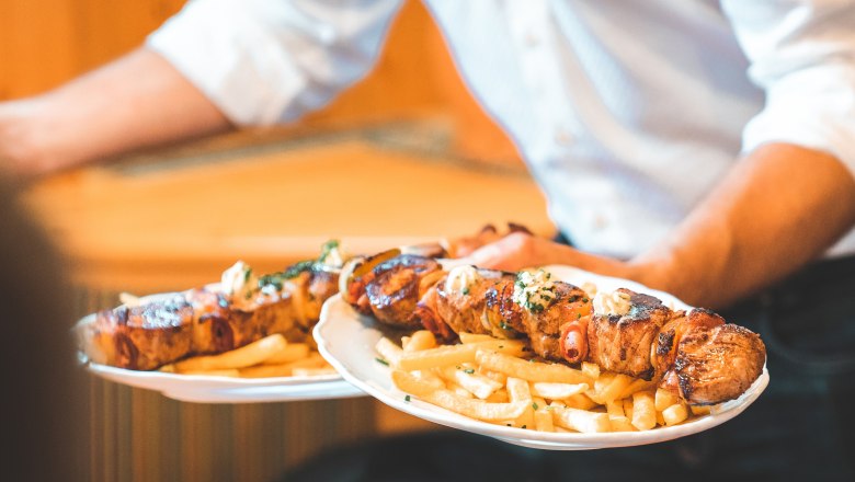 Hubertushof, © Matthias Auer A waiter serves two plates of grilled meat skewers and chips.