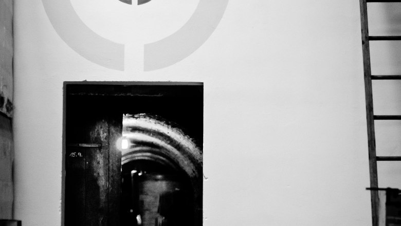 Obenaus-Keller, © Bernhard Kastner A black and white photo of a cellar with a chair in front of a door and a target pattern on the wall.