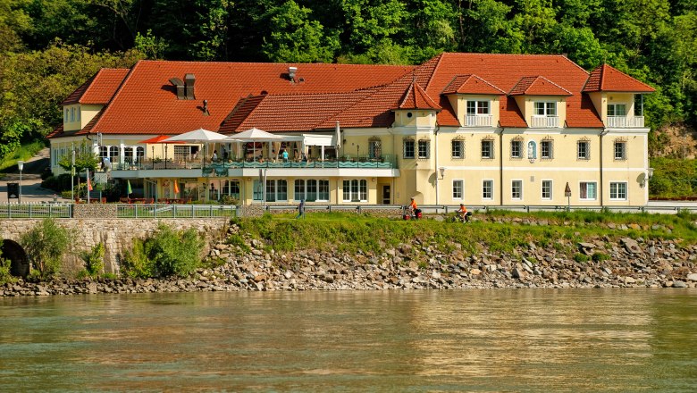 Residence Wachau, © m+m webeagentur A yellow building with red roofs on the riverbank, surrounded by trees.