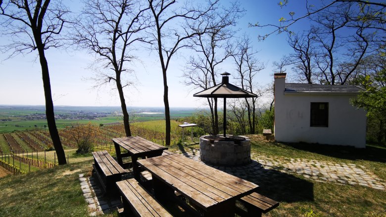Linger on the Hutberg, © Weinstraße Weinviertel Picnic area with wooden tables and barbecue on a hill overlooking vineyards and a small building.