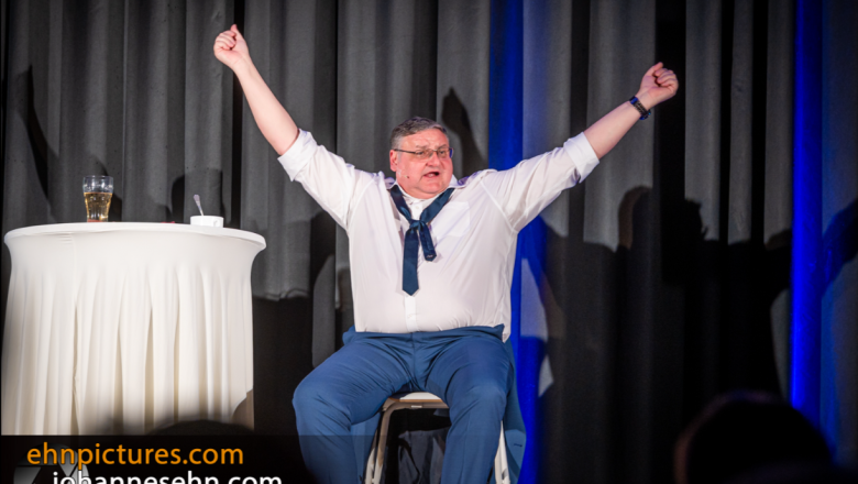 Stage, cabaret artist with glasses white shirt blue pants, atmospheric, laughing,, © Gasthubers Wirtshaus A man sits on a stage with his arms raised, next to him a table with a glass.