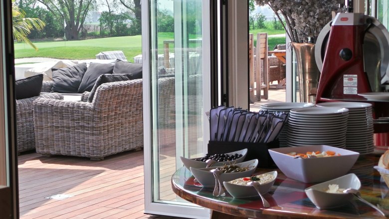 Boathouse, © DCC A laid table with crockery and snacks, next to a terrace with rattan furniture and a view of a green lawn.