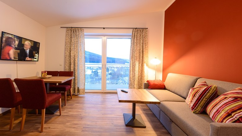 Heart stone room, © Wirtshausbrennerei Krenn, Johann Frank Cozy living room with red accent wall, grey sofa, wooden table, TV and large window with curtains.
