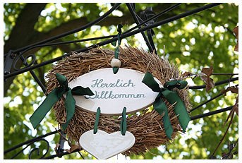 Welcome sign, © Restaurant Krone in Gaaden A welcome sign in a wreath with green ribbons, hung on a metal frame, against a background of green leaves.