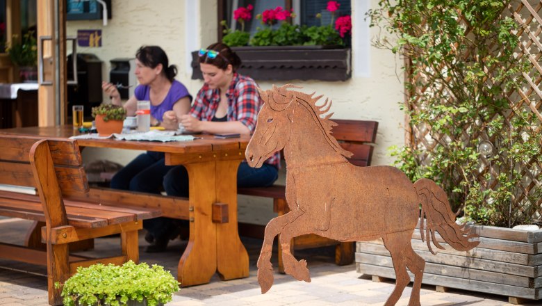 Schagerlhof Joachimsberg, © Fred Lindmoser Two women sit at a wooden table outdoors and eat. In the foreground is a metal sculpture of a horse.