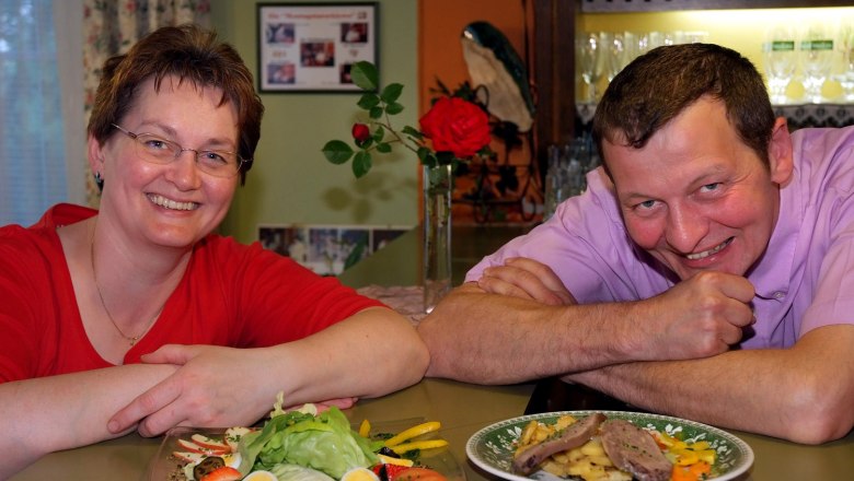 Autumn family, © Seymannfilm A man and a woman smile at the camera, with plates of food in front of them. A red rose and a shelf with glasses can be seen in the background.