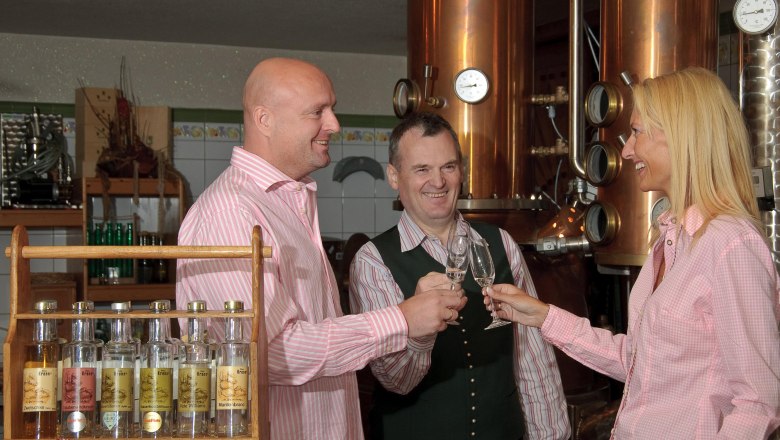 Krenn Distillery, © Wirtshausbrennerei Krenn Three people clink glasses of schnapps in a distillery.