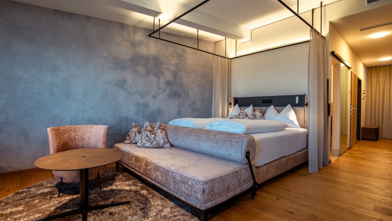 Hotel Schachner, © Niederösterreich Werbung / Maximilian Pawlikowsky Modern hotel room with four-poster bed, armchair and table on wooden floor.