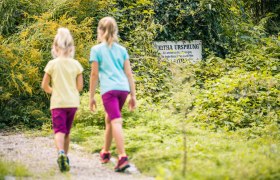 In the Leitha-Au, © Wiener Alpen, Martin Fülöp Two children are walking along a path in a green wooded area. An ivy-covered sign with the inscription 'Leitha Ursprung' is visible in the background.