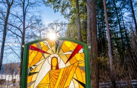 Glass Way of the Cross, © Wiener Alpen, Flotoanker - Luckerbauer Stained glass window in the forest with sunshine in the background.