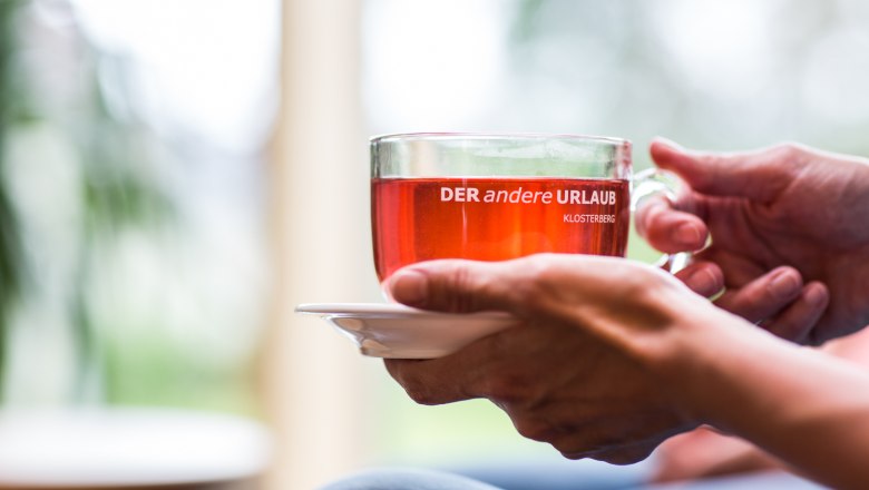 Fasting, © Gesundheitshotel Klosterberg GmbH Close-up of hands holding a cup with a red drink on which is written 'DER andere URLAUB KLOSTERBERG'.