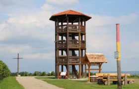 Observation tower in Roseldorf, © Gemeinde Sitzendorf Wooden lookout tower with red roof on a meadow, next to it a wooden cross and a bench.