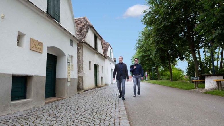 In the wine cellar lane, © Elke Wolfbeisser Two men walk along a cobbled street, past white buildings and trees.
