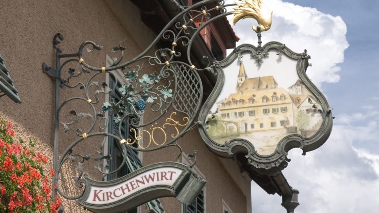 Kirchenwirt, © Kirchenwirt An artistically designed inn sign with the inscription 'Kirchenwirt' and a picture of a building with a church tower.