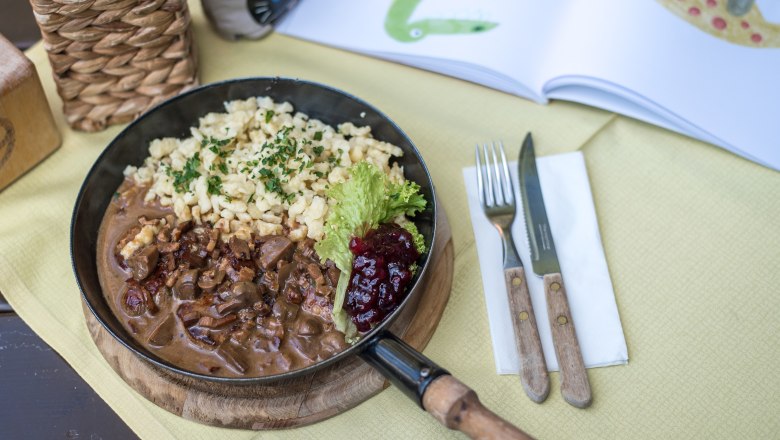 Monthly seasonally changing dishes, © Niederösterreich Werbung/Claudia Schlager A plate of spaetzle, meat and cranberries on a table with cutlery and an open book.