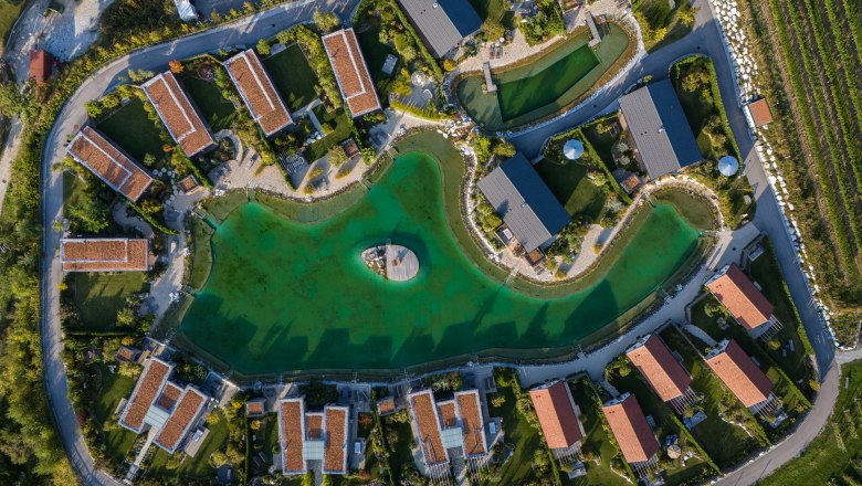 Kittenberger Chalets at the Gartensee, © Niederösterreich Werbung / Maximilian Pawlikowsky Aerial view of chalets around a green lake with a jetty in the middle.