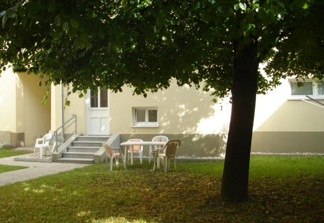 Garden, Apartment Gutmann, © Gutmann Inner courtyard with lawn, a tree and white plastic chairs. The yellow building in the background.