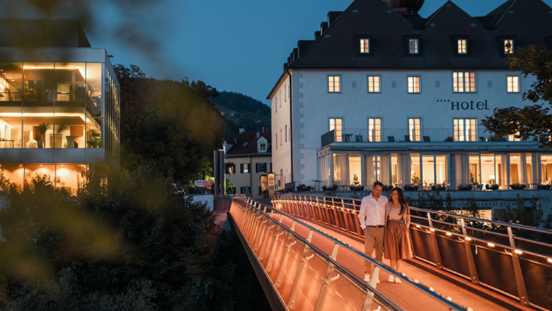 The castle on the Iron Road, © Dominik Stixenberger A couple walks across an illuminated bridge in front of a hotel in the evening.