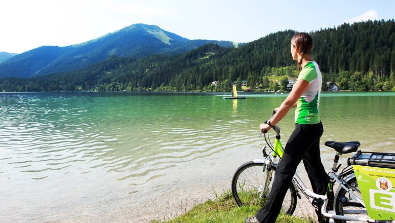 Turquoise blue water at Lake Erlaufsee, © weinfranz.at Woman with bicycle on the shore of Lake Erlauf, surrounded by mountains and forest.