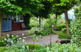 Show garden Am Pulverturm, garden house, © Schaugarten Am Pulverturm An idyllic garden with a small wooden house, surrounded by trees and plants, with a gravel path and a small pond.