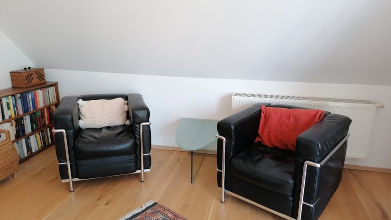 Living area in the "balcony room", © Wiener Alpen Two black leather armchairs with cushions, a glass side table, bookshelf and carpet in a living area with a sloping roof.