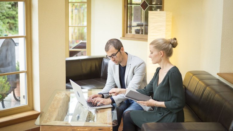 Alpen-Aktiv-Landgasthof zur Schubertlinde, © Patrick Haberler Two people work on a laptop in a cozy room with large windows.