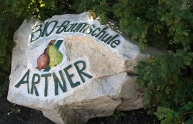 Artner Organic Nursery, © Bio-Baumschule Artner Large stone with the inscription 'Bio-Baumschule Artner' and painted fruit, surrounded by green plants.
