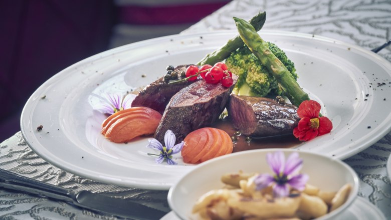 Saddle of venison with pepper apricots, broccoli, potato noodles, © Niederösterreich Werbung/Andreas Hofer A plate of saddle of venison, peppery apricots, broccoli and asparagus, decorated with flowers. In the foreground, a bowl of potato noodles.
