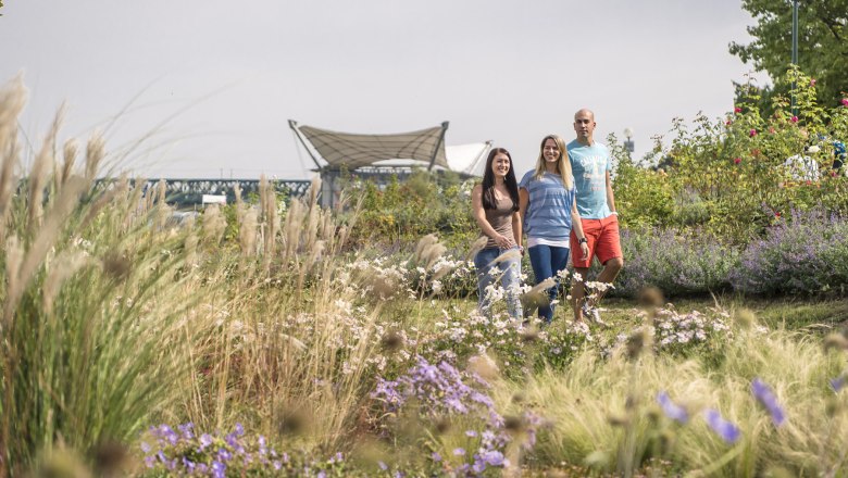donaulaende1_c_robert-herbst-stadtgemeinde-tulln, © Robert Herbst/Stadtgemeinde Tulln Three people stroll through a blooming garden with grasses and flowers, a bridge and a modern roof in the background.