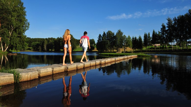 Schrems mud bath, © Waldviertel Tourismus, Robert Herbst Two people walk on a footbridge over a lake in the Schrems moor spa, surrounded by trees and blue sky.