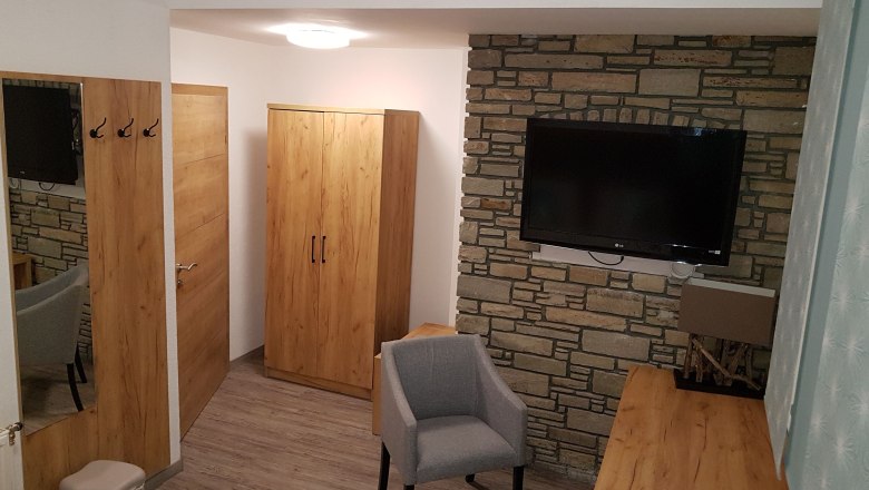 Pension Central, © Konrad Brandl Hotel room with wooden furniture, gray armchair and flat screen TV on a stone wall.