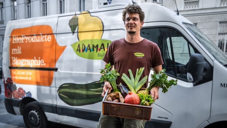 ADAMAH BioKistl, © Thomas Apolt A man holds a box of fresh fruit and vegetables in front of a delivery van with the ADAMAH logo.