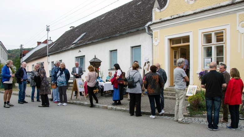 Hardegg watercolor painting group, © Rosi Grieder A group of people are standing in front of a yellow building on a street, chatting. Some of them are looking at works of art on display outside.
