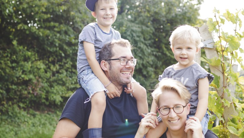 TDR family photo, © Buschenschank Buder Family outdoors, two adults carrying children on their shoulders, sunny day.