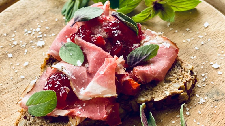 Wild smoked meat, © Amelie Seilern-Aspang Close-up of a sandwich with ham, cranberries and herbs on a wooden board.