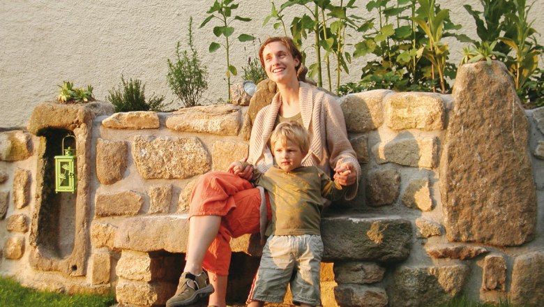 Prinzenhof, © Prinzenhof A woman and a child sit on a stone bench in front of a rock garden overgrown with plants.