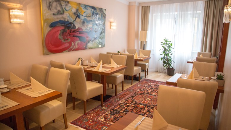 Breakfast room, © Hotel Admiral am Kurpark An elegant breakfast room with laid tables, beige chairs, a colorful carpet and a large abstract painting on the wall.