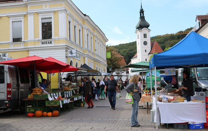 Farmers' market, © Wienerwald Tourismus GmbH A busy farmers' market with stalls and visitors in front of a yellow building and a church in the background.